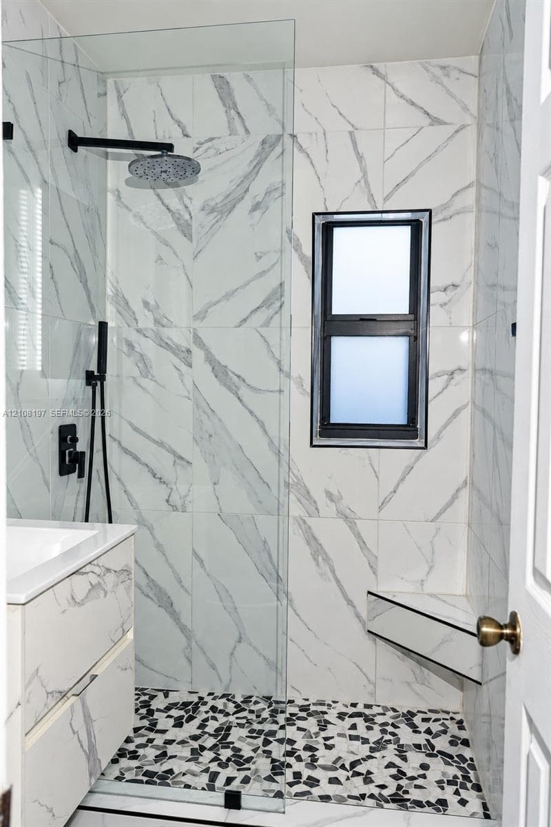 Walk-in shower featuring large-format stone-patterned wall tile, a built-in corner seat, matte black rainfall showerhead with handheld attachment, pebble-texture flooring, and a frameless glass partition