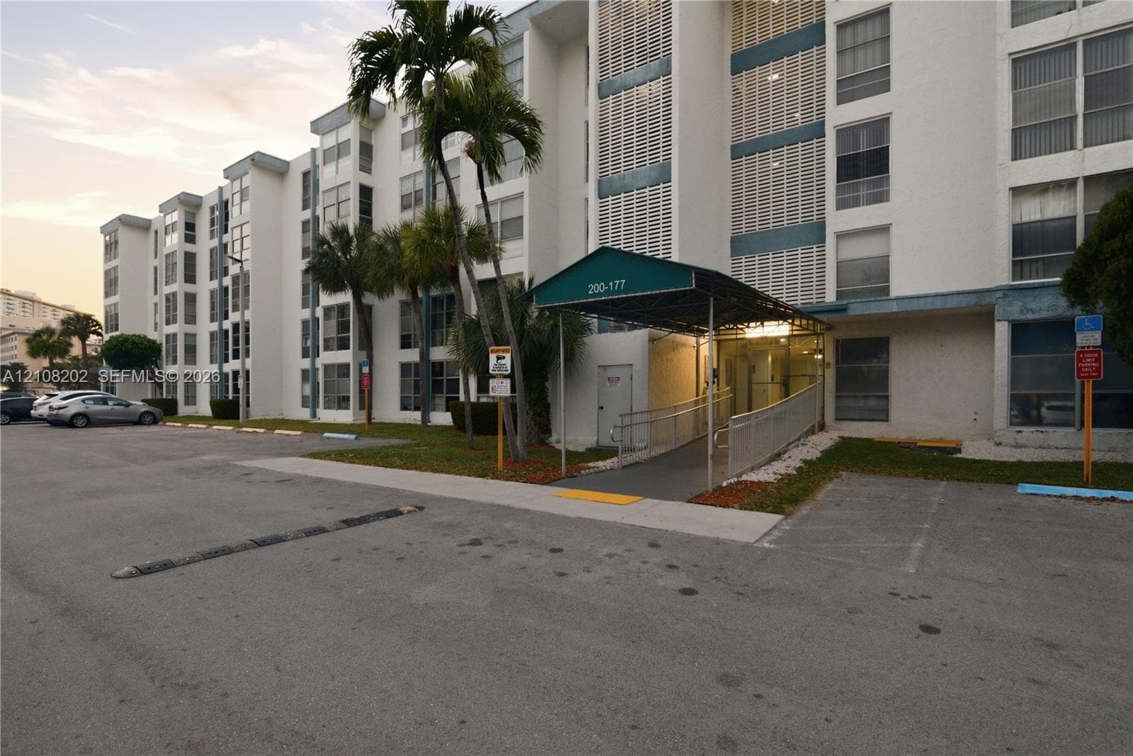 Multi-story exterior featuring a concrete facade, covered entryway with a metal awning, paved surface parking, and mature palm trees