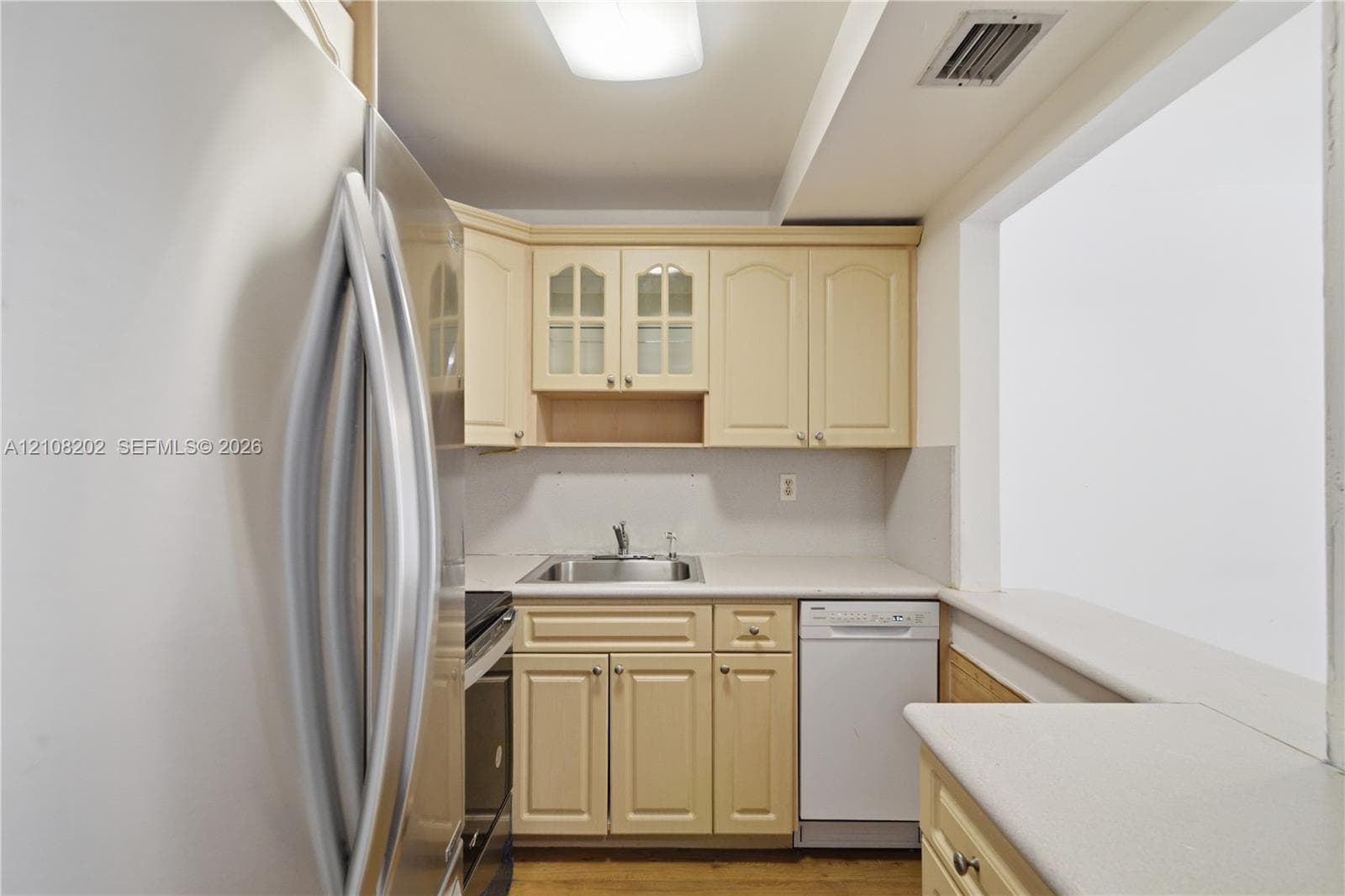 Kitchen featuring light-tone cabinetry with glass-paneled upper doors, solid-surface countertops, an integrated stainless steel sink, and a built-in dishwasher