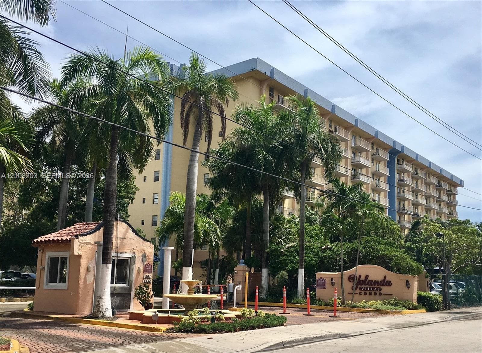 Multi-story residential complex featuring a gated entrance, a decorative tiered fountain, mature palm trees, and a paved brick driveway