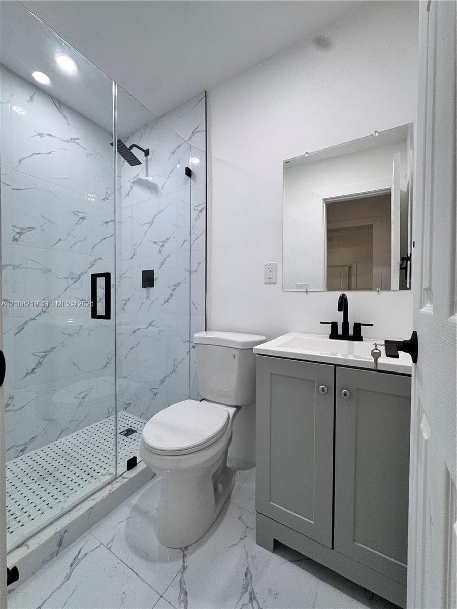 Walk-in shower with stone-finish tile surround and frameless glass door, gray Shaker-style vanity, matte black plumbing fixtures, and stone-finish floor tiling
