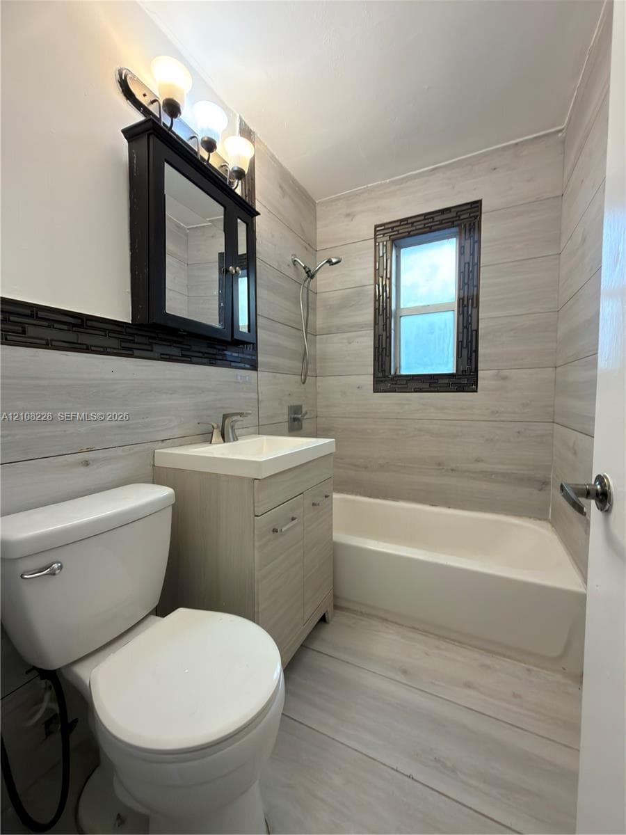 Bathroom featuring light-toned wood-finish wall and floor tile, a recessed medicine cabinet with integrated lighting, a modern vanity with an inset sink, and a tiled bathtub surround with a mosaic accent border