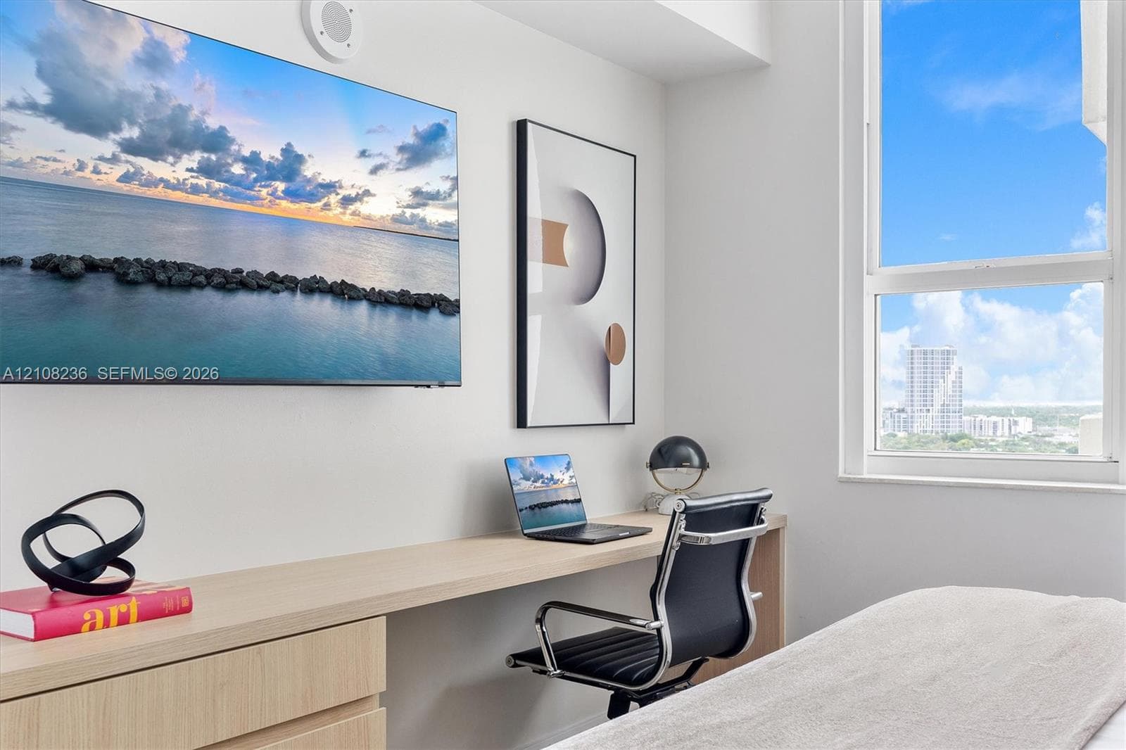Custom built-in desk with light wood-finish cabinetry, ceiling-mounted speaker, and large-format window featuring expansive sky and urban scenery