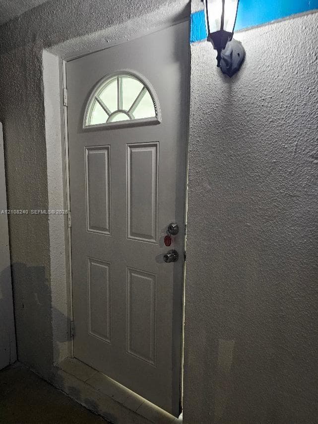 Exterior entry door featuring a semi-circular transom window, raised-panel design, and a wall-mounted lantern-style light fixture against a textured stucco exterior finish