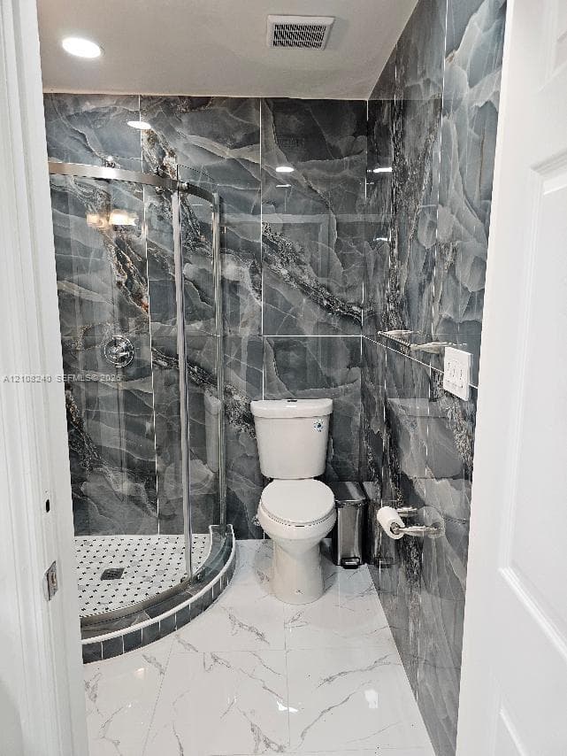 Bathroom featuring floor-to-ceiling dark-veined wall tile, a corner glass-enclosed shower with a mosaic-tile basin, and large-format white tile flooring with veining detail