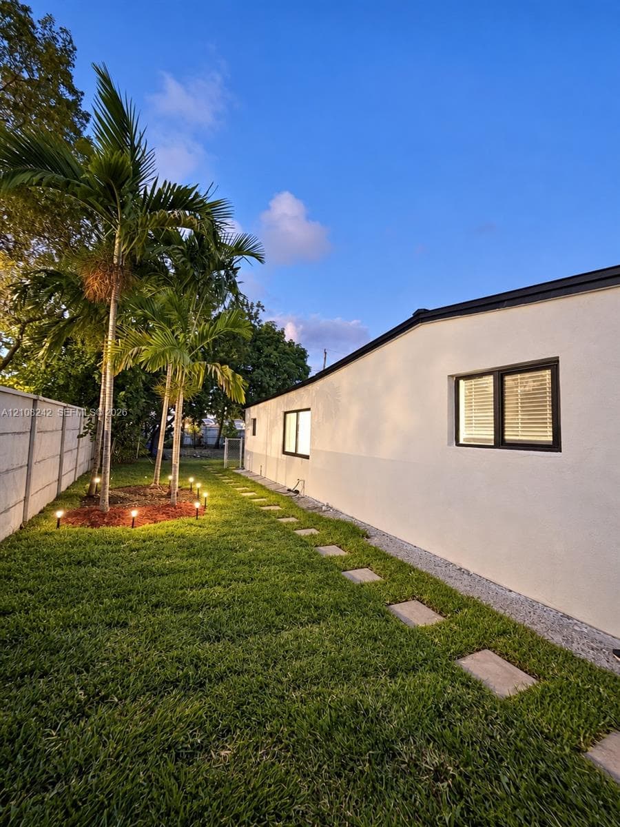 Side yard featuring a manicured lawn, established palm trees, a concrete block privacy wall, stone paver pathway, and recessed landscape lighting