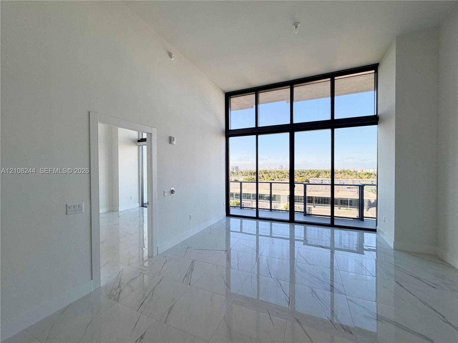 High-ceilinged interior featuring floor-to-ceiling windows with black metal frames, polished stone-finish tile flooring, and a glass-paneled balcony