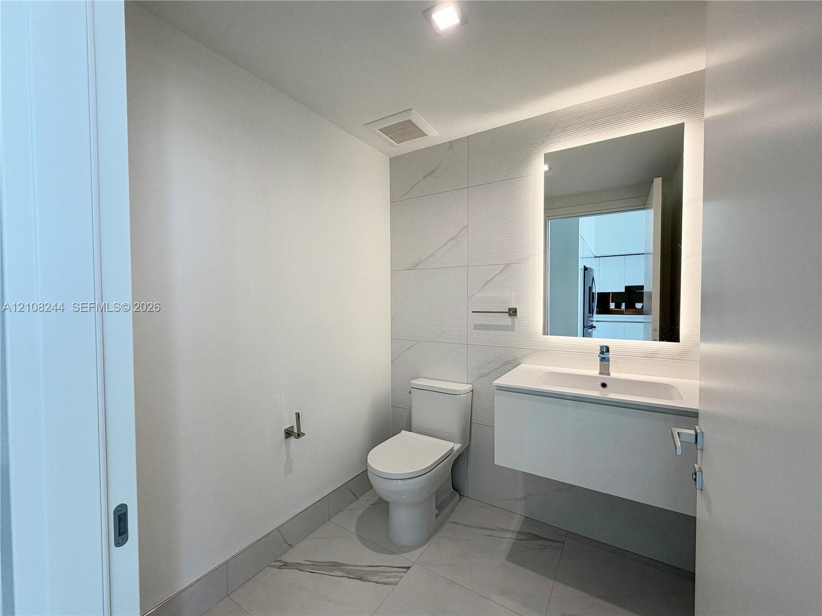 Contemporary bathroom featuring a floating white vanity, polished large-format tile flooring, wall-to-wall decorative tile backsplash, and a modern backlit mirror