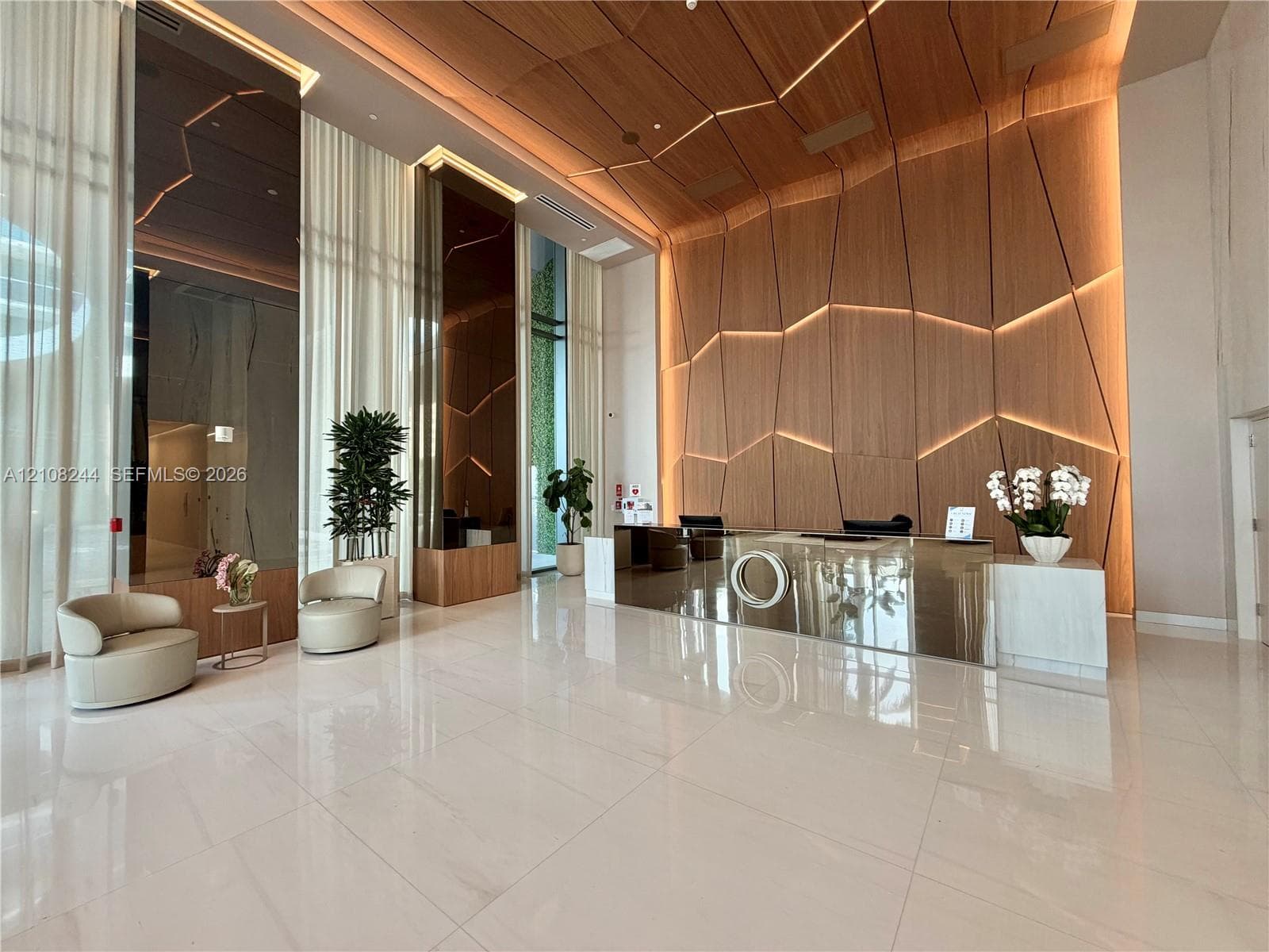 Lobby featuring high-gloss tile flooring, floor-to-ceiling windows with full-length drapery, geometric wood-paneled walls with integrated accent lighting, and a metallic reception desk