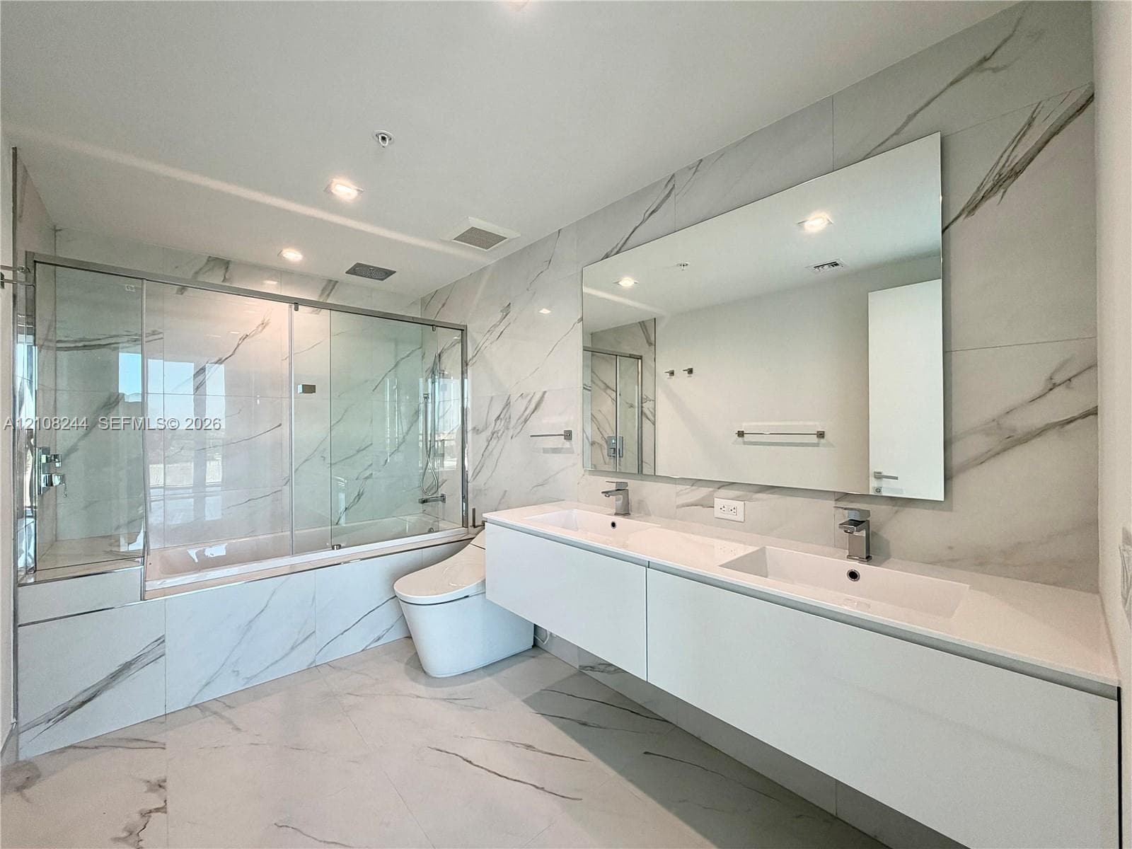Floating double vanity with integrated sinks, full-wall mirror, glass-enclosed soaking tub with shower combination, and large-format porcelain tile wall cladding