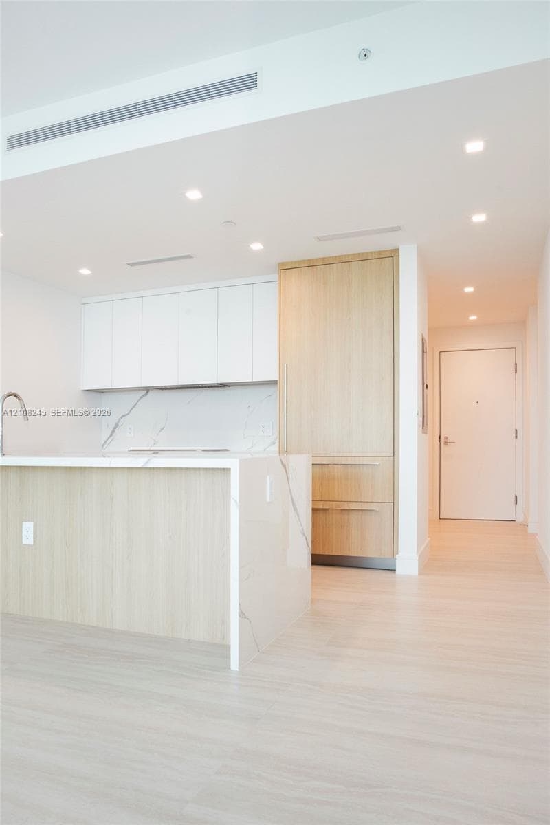 Kitchen featuring waterfall-edge stone-finish island, light wood-finish cabinetry, white flat-panel upper cabinets, integrated appliances, and recessed ceiling lighting