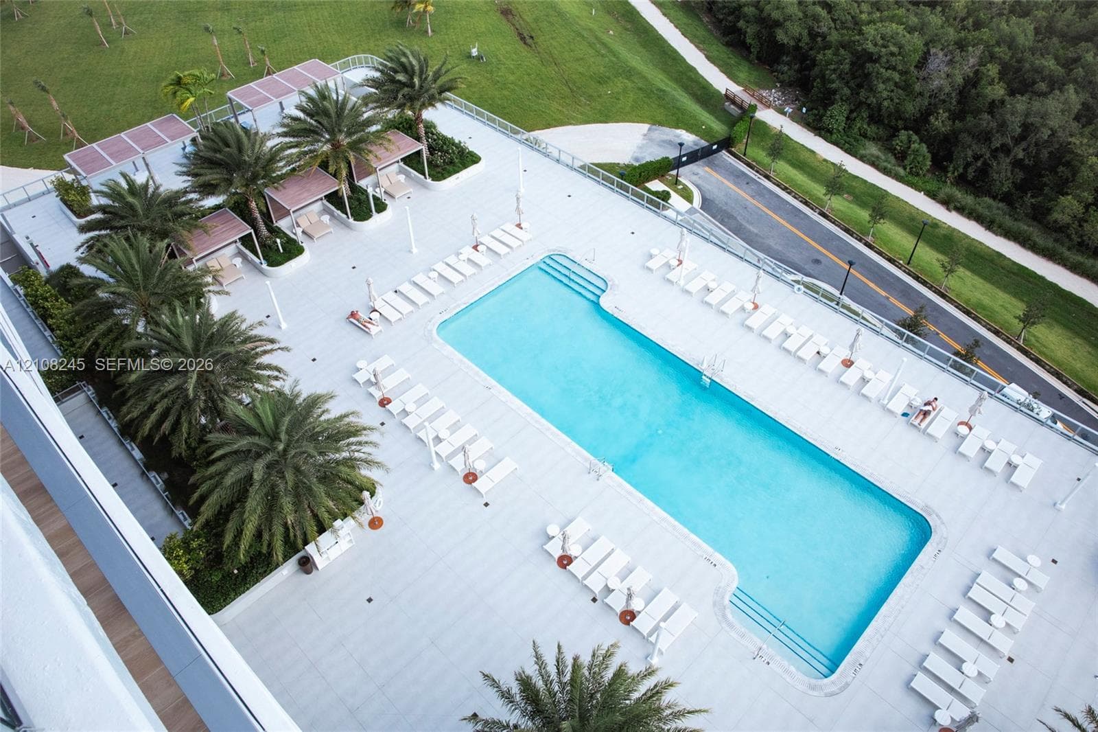 Rectangular swimming pool with stepped entry, surrounded by a paved deck featuring shaded cabanas, palm landscaping, and perimeter metal railings