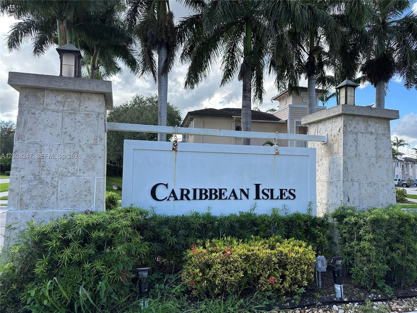 Community entrance featuring dual stone-clad pillars, attached lanterns, and mature palm trees