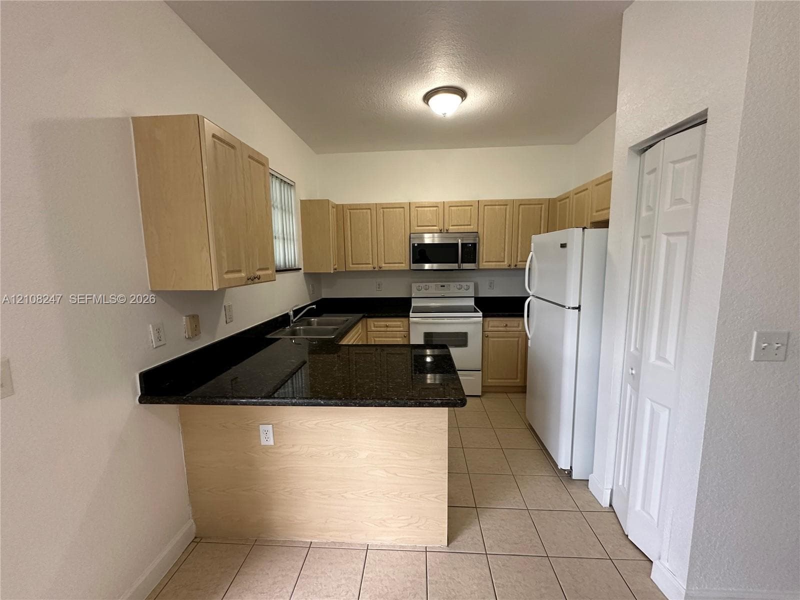 Kitchen featuring light-finish cabinetry, dark solid-surface countertops, tile flooring, and integrated white appliances