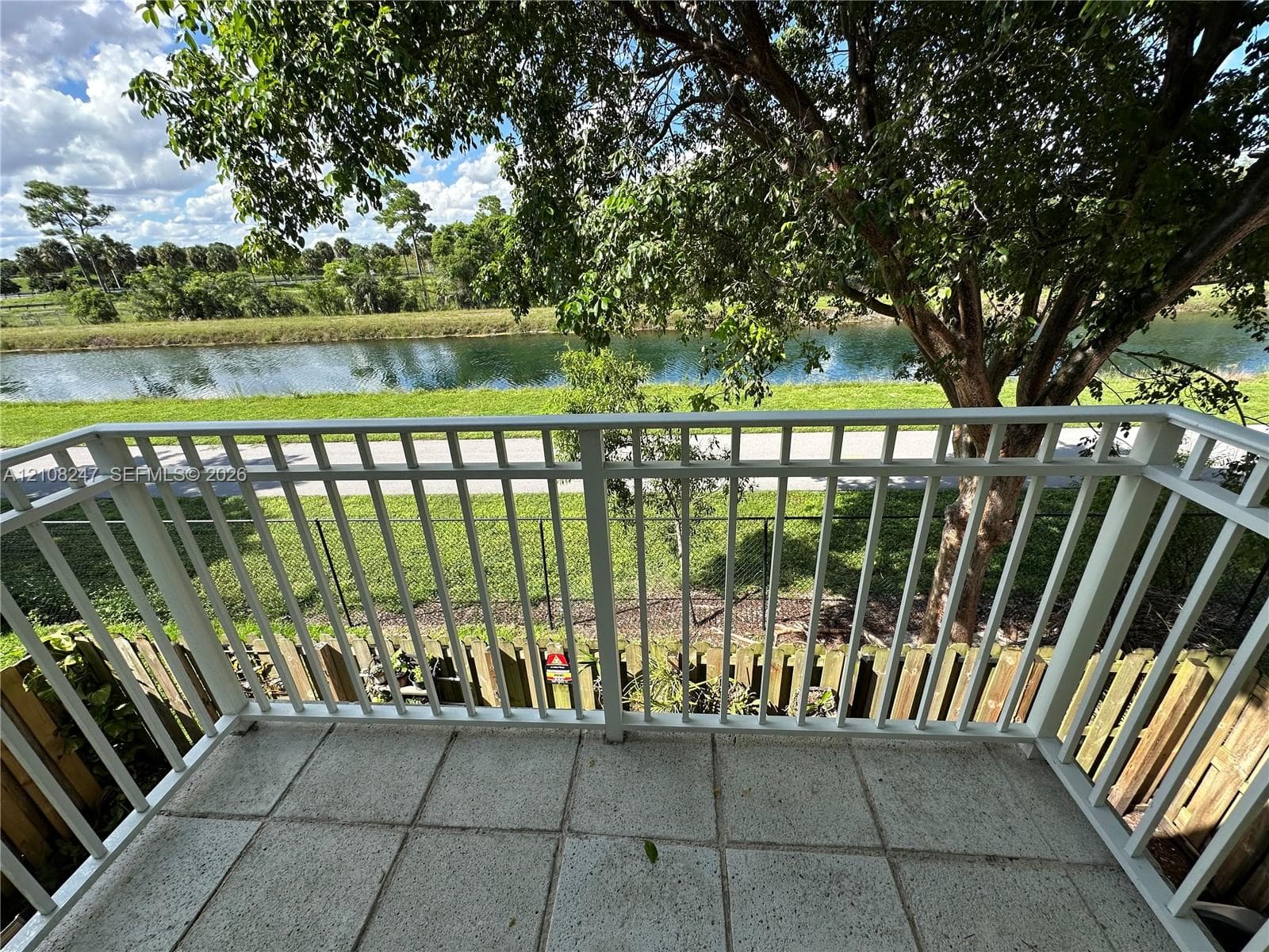 Balcony featuring white aluminum railings, light-toned concrete tile flooring, and a backdrop of mature trees overlooking a canal