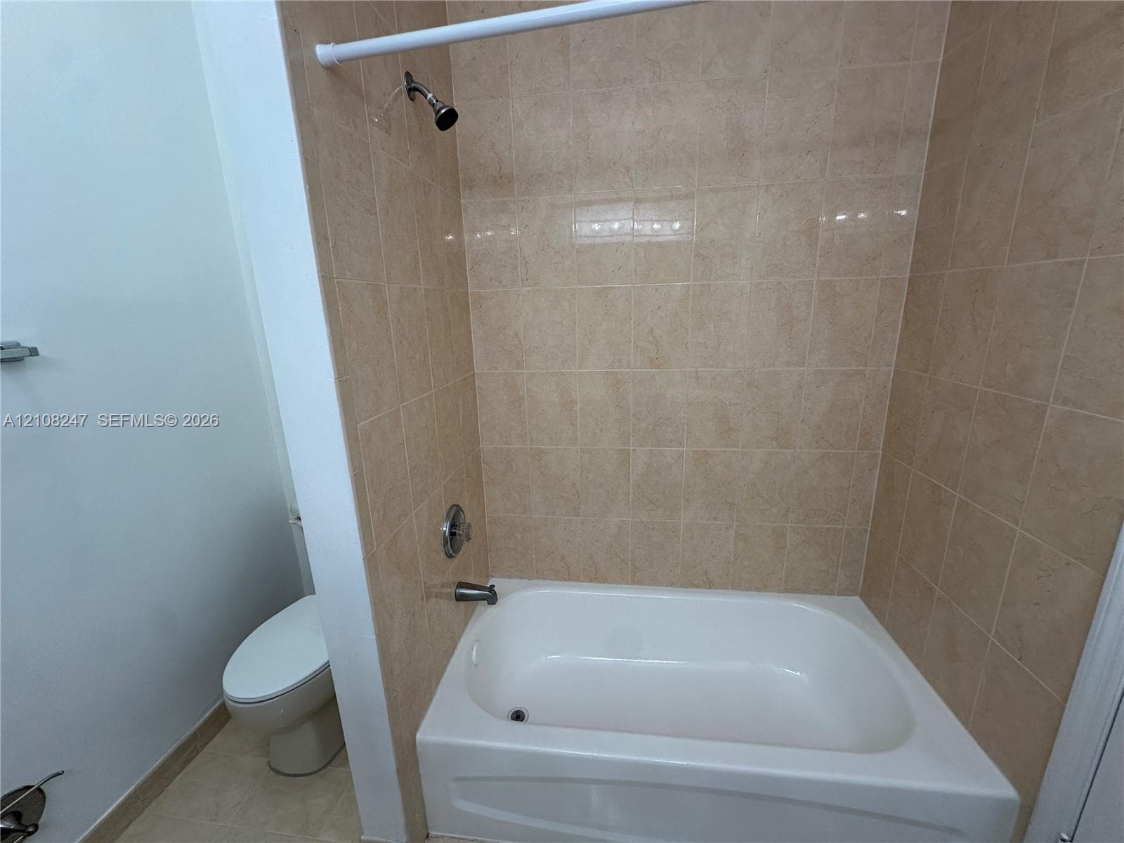 Bathtub with a white acrylic surround, tan wall-tile finish, showerhead, and a standard-height toilet