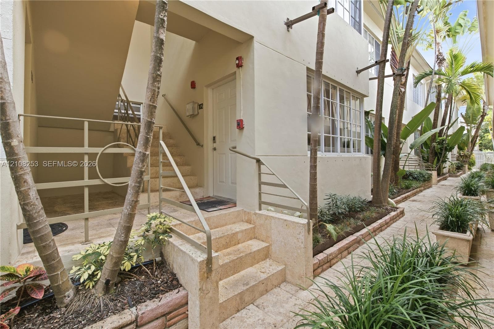 Exterior walkway featuring natural stone pavers, concrete stairs, metal handrails, and built-in landscape planters with mature palms and shrubbery