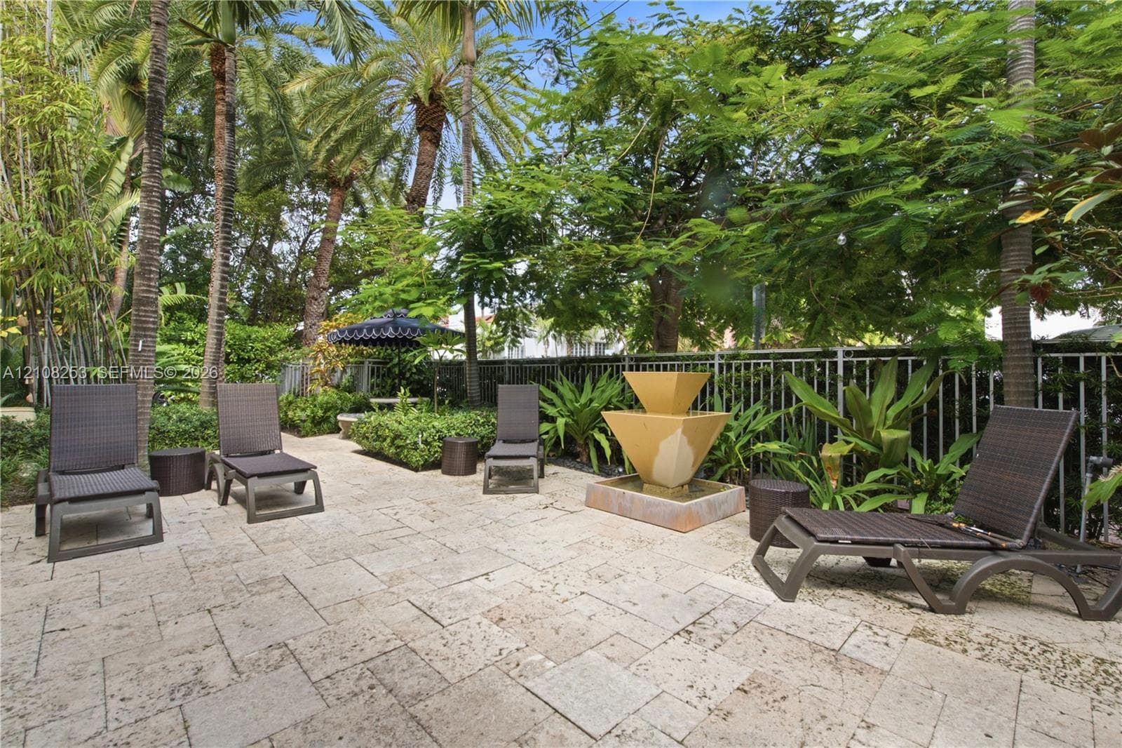 Stone paver patio featuring a tiered fountain, metal perimeter fencing, and mature landscaping