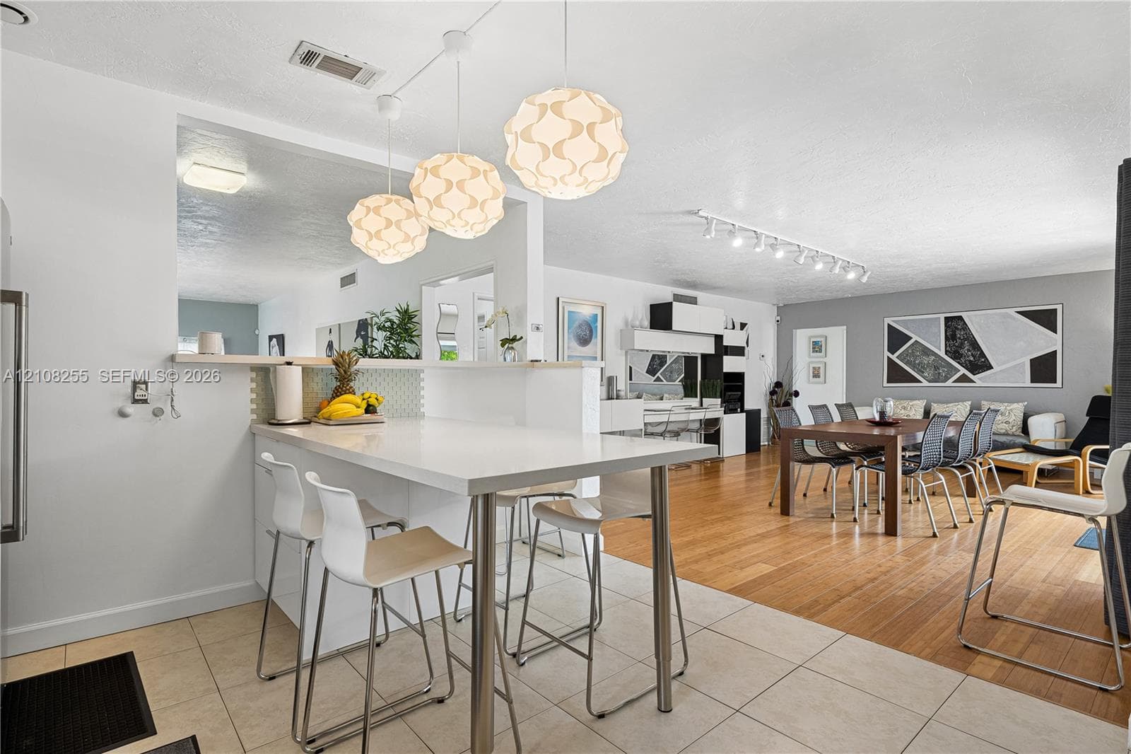 Kitchen peninsula with a solid-surface countertop and integrated breakfast bar, complemented by three spherical pendant light fixtures, large-format tile flooring, and wood-finish flooring