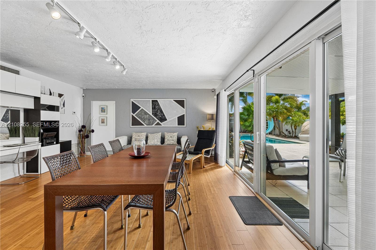 Living area featuring wood-finish flooring, built-in cabinetry, ceiling-mounted track lighting, and expansive sliding glass doors providing access to a paved patio and outdoor swimming pool