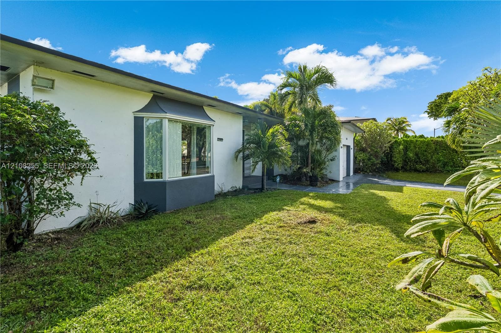 Single-story exterior featuring white stucco siding, a charcoal-trimmed bay window, mature palms, and expansive lawn