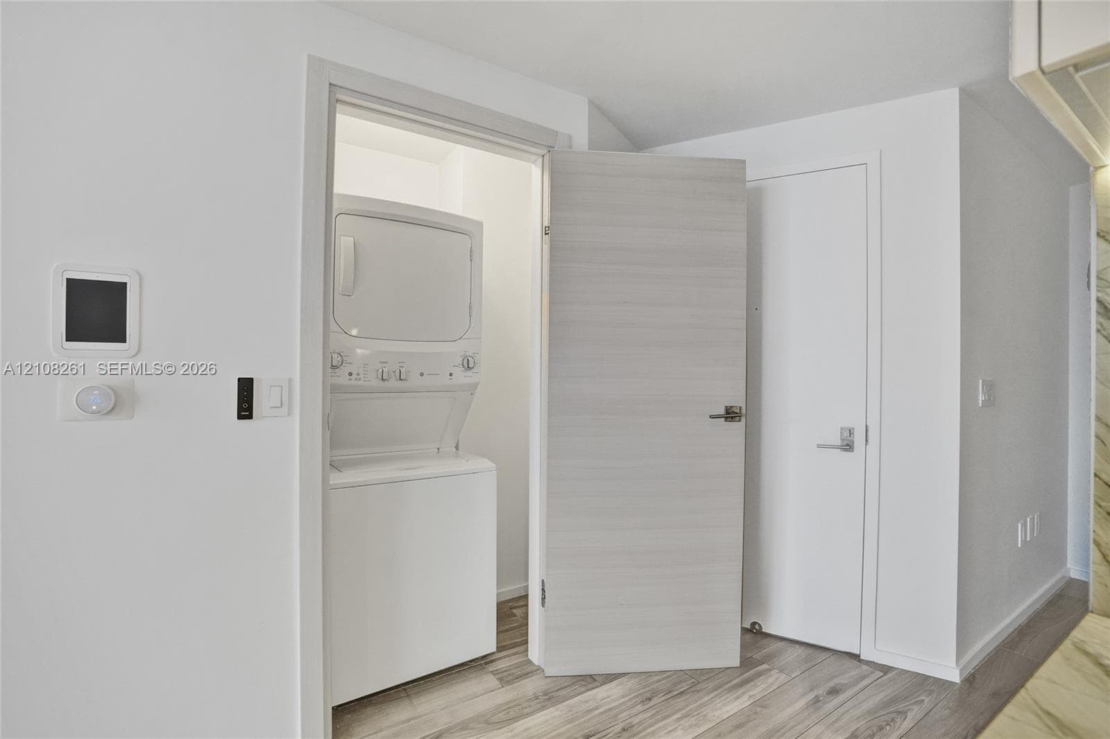 Laundry closet featuring a stackable washer and dryer, light wood-finish door, wood-finish flooring, wall-mounted smart home controls, and interior doors with lever-style handles