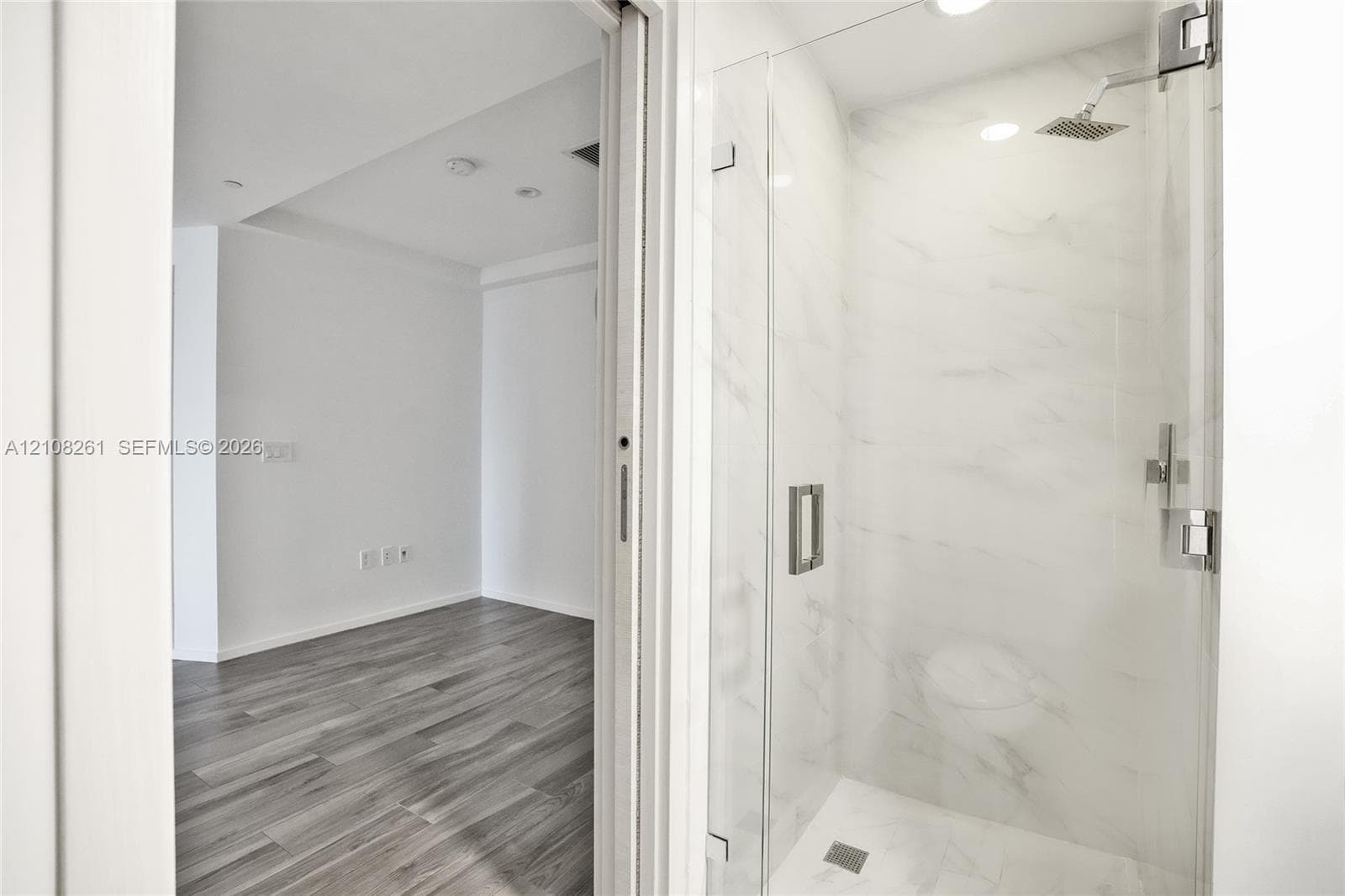 Walk-in shower featuring a glass enclosure, wall-mounted rain shower head, and full-height stone-finish tile surround, complemented by wood-finish flooring in the surrounding space