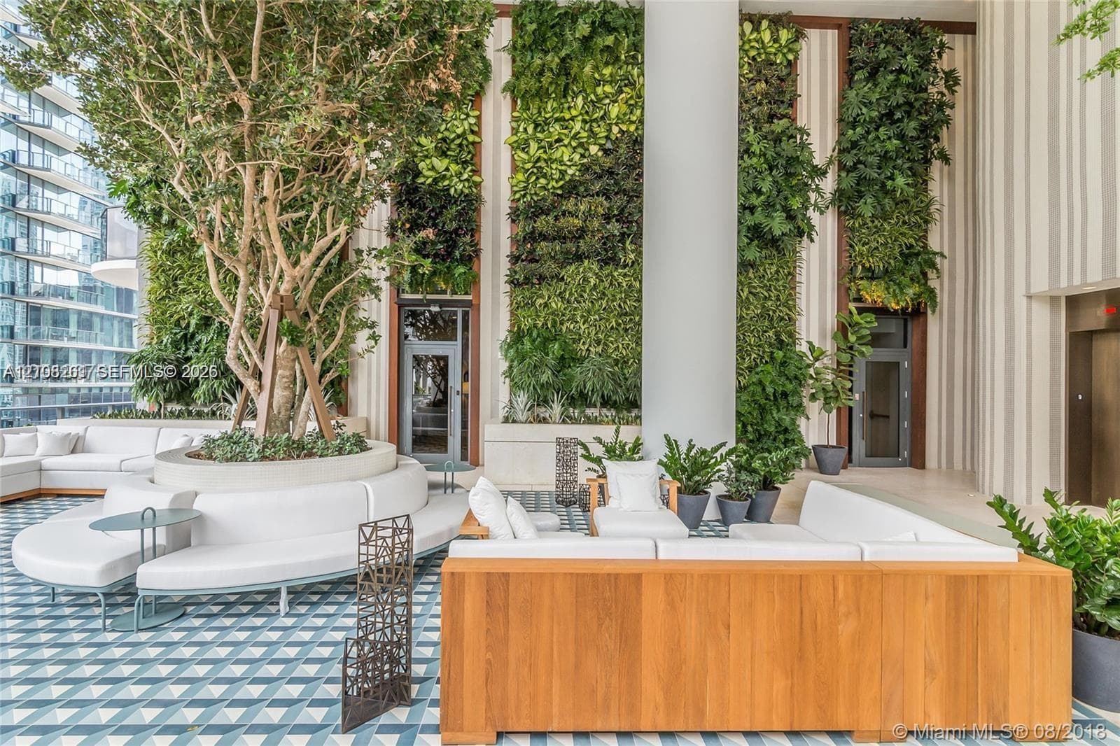 Covered lounge area featuring a geometric patterned tile floor, full-height vertical gardens, a mature tree enclosed by a circular planter, and a clean, neutral-toned structural column