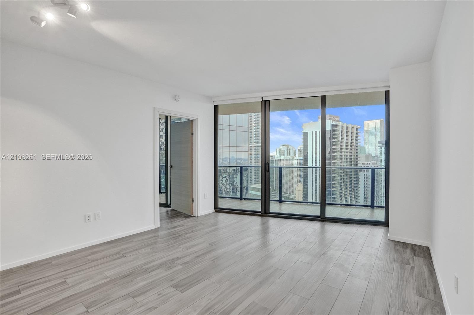 Interior room featuring wood-finish flooring, floor-to-ceiling sliding glass doors, a private balcony with glass railing, and urban skyline surroundings