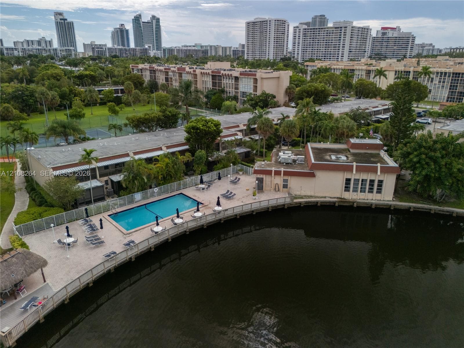 Waterfront swimming pool with a paved deck, perimeter fencing, and a thatched-roof cabana overlooking a canal