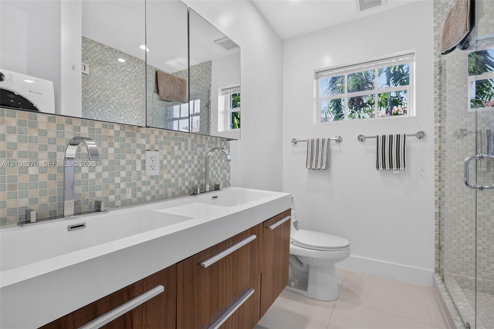 Dual-sink floating vanity with wood-finish cabinetry, mosaic tile backsplash, mirrored medicine cabinets, and glass-enclosed shower