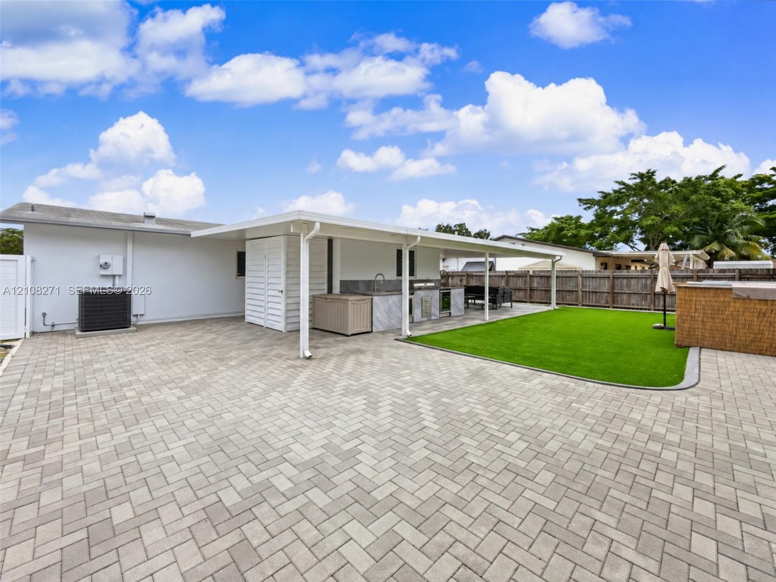 Rear exterior featuring a covered patio, paved hardscaping, synthetic turf area, and built-in outdoor kitchen with stone-finish cabinetry
