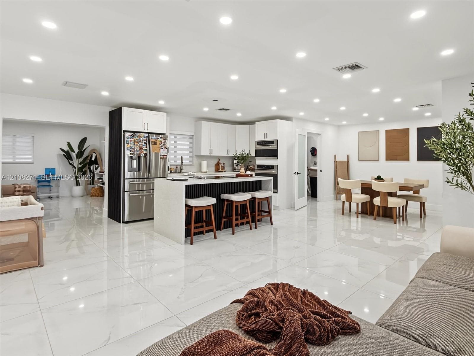 Large-format polished tile flooring, central kitchen island with waterfall-edge countertop and fluted detail, white cabinetry, integrated stainless steel appliances, and recessed lighting