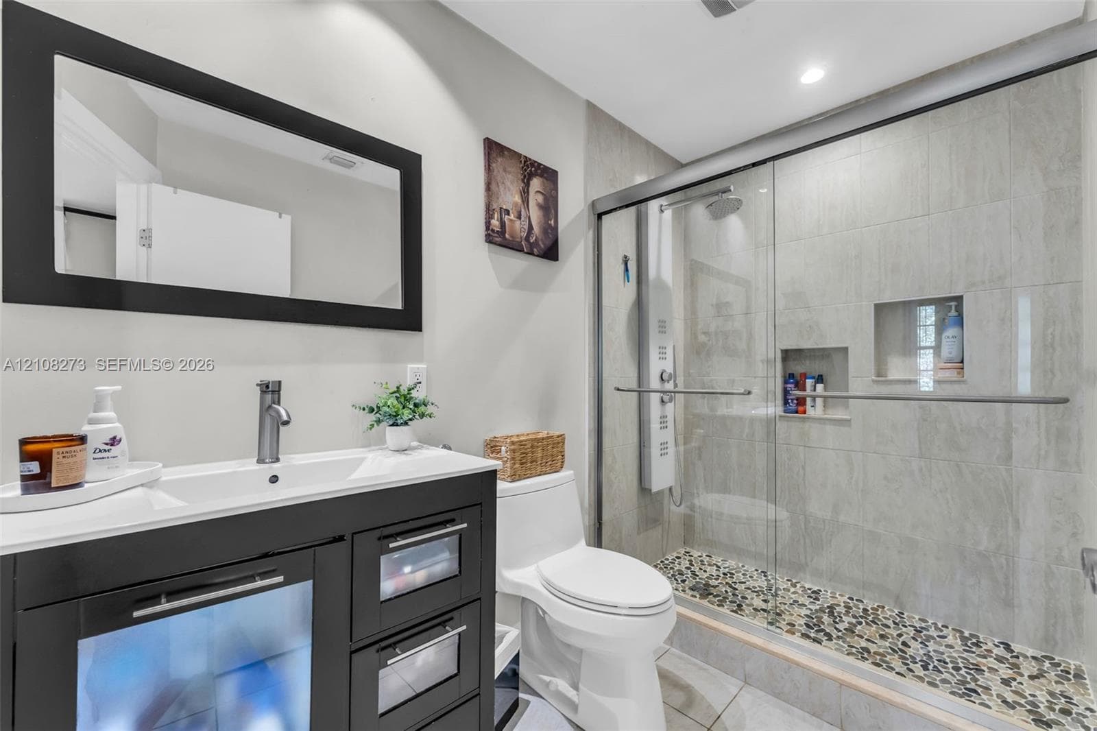 Bathroom featuring a dark wood-finish vanity with glass-panel doors, wall-mounted mirror with a dark frame, walk-in shower with a glass enclosure, pebble-tile shower flooring, and built-in shower shelving