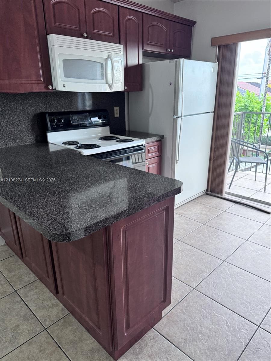 Kitchen featuring dark toned cabinetry, ceramic tile flooring, and white appliances, with sliding door access to an exterior balcony