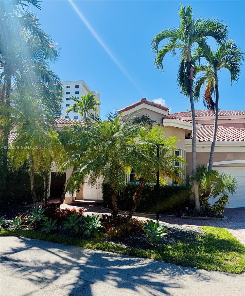 Stucco exterior featuring a terracotta tile roof, paver driveway, attached garage, and mature palm landscaping