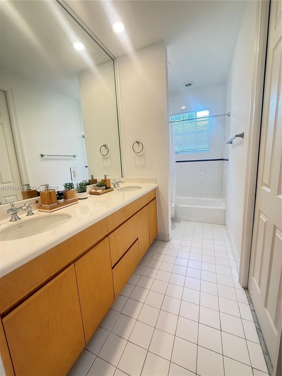 Dual-sink vanity with light wood-finish cabinetry, solid surface countertop, expansive wall-mounted mirror, and tile flooring