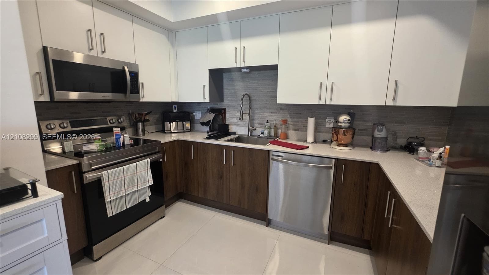 Kitchen featuring two-tone flat-panel cabinetry, light-toned stone-finish countertops, stainless steel appliances, a textured tile backsplash, and an undermount sink with a pull-down faucet