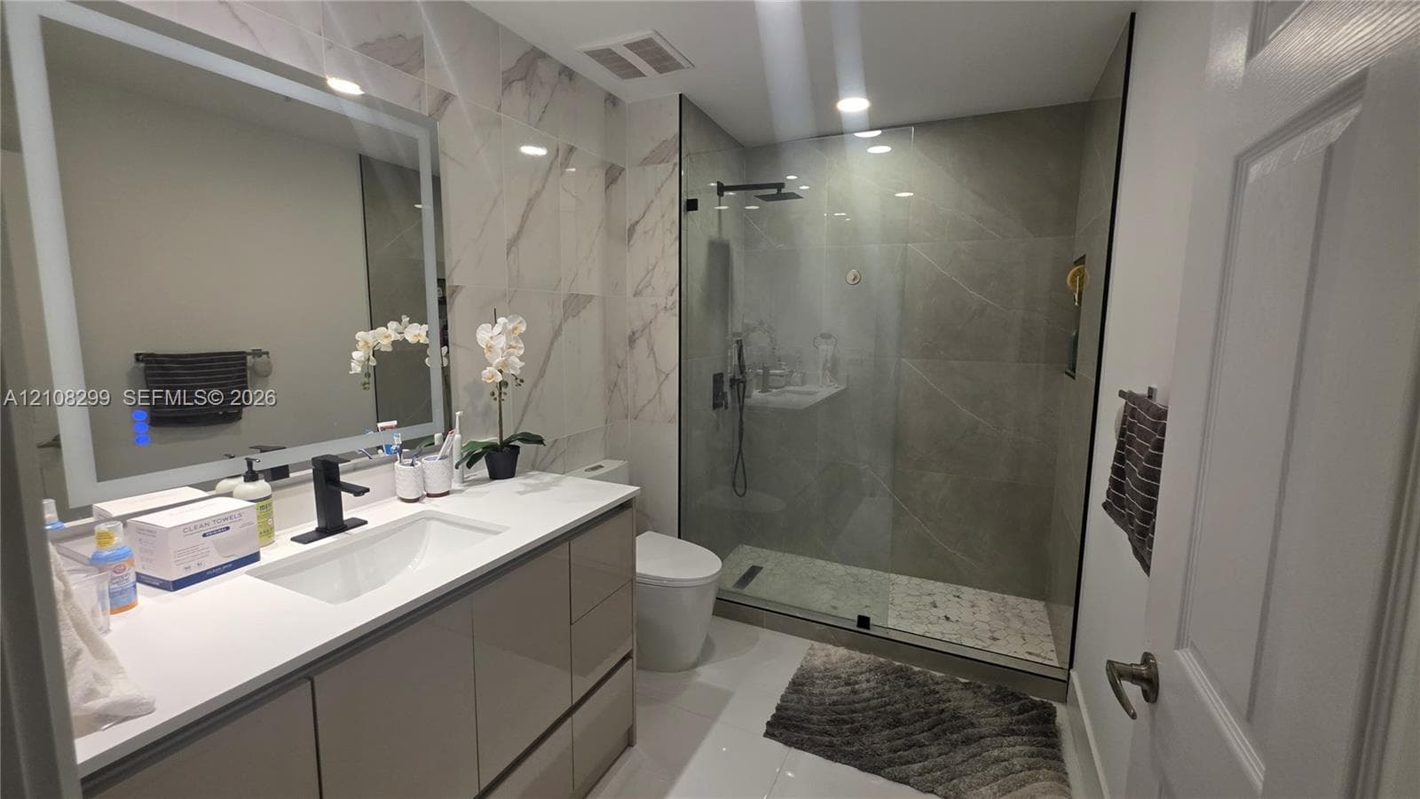Modern bathroom featuring a high-gloss vanity with a white countertop, wall-mounted illuminated mirror, and a walk-in shower with a glass enclosure, rainfall showerhead, and stone-finish floor tiling