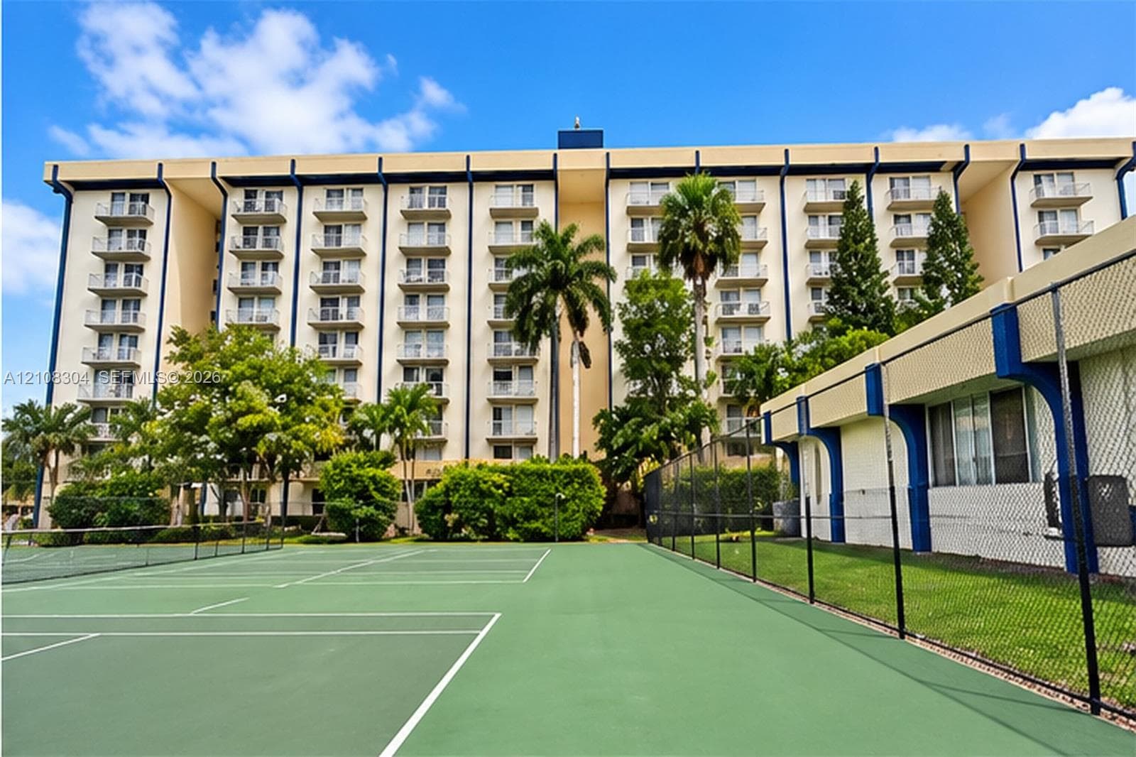Multi-story residential building featuring private balconies, tennis courts, chain-link perimeter fencing, and established palm trees