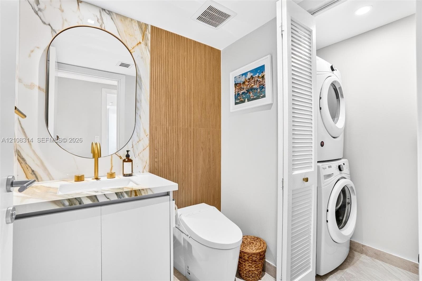 Stone-surround vanity with a circular mirror and gold-tone faucet, complemented by vertical slat wood-finish wall paneling, a low-profile toilet, and a louvered-door closet containing a stacked laundry unit