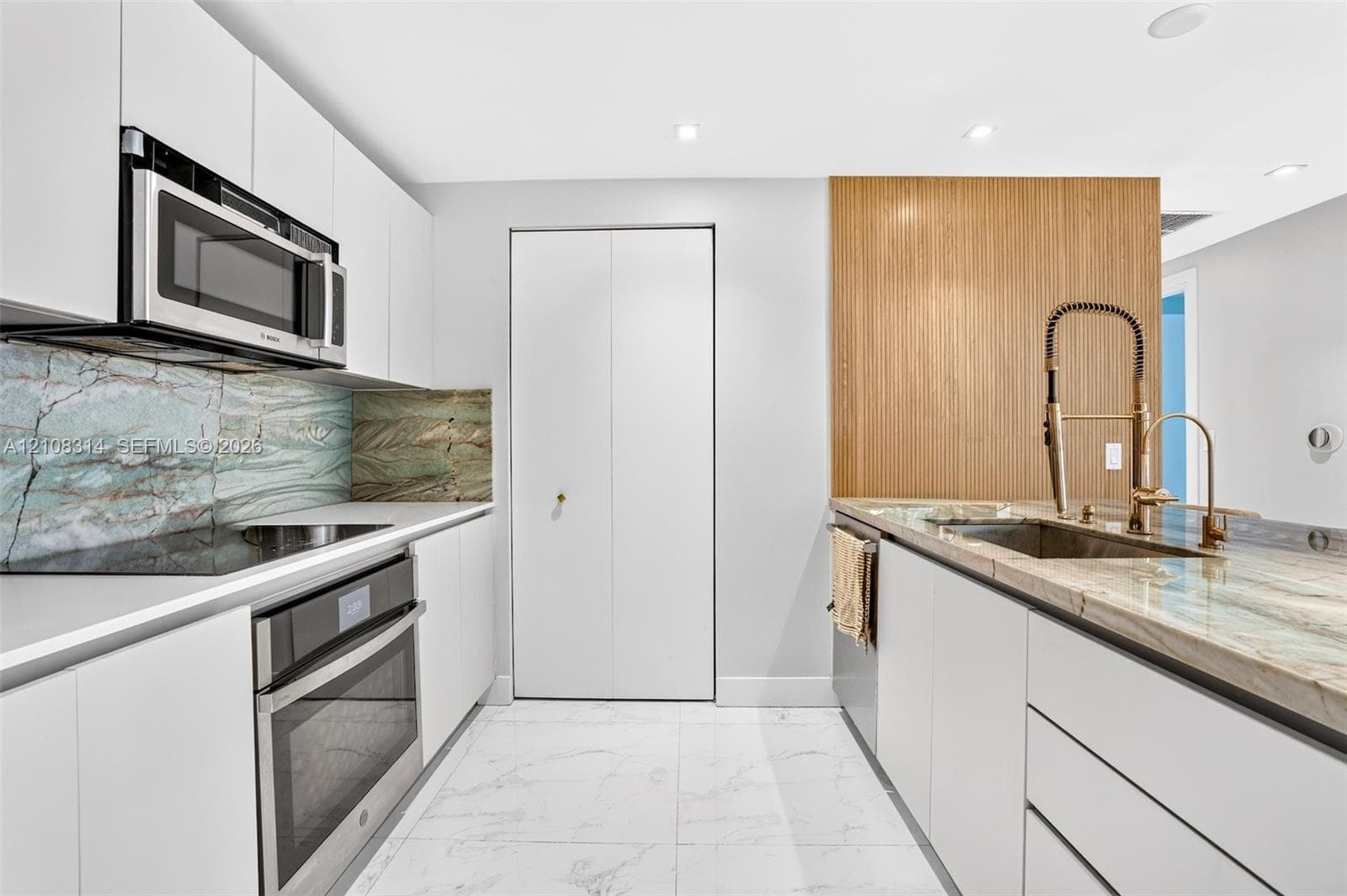 Modern kitchen featuring flat-panel white cabinetry, stone slab countertops, integrated stainless steel appliances, and wood-finish accent paneling with a gooseneck faucet