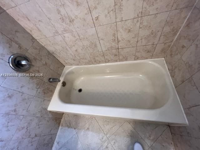Bathtub enclosed by floor-to-ceiling ceramic wall tile and featuring a chrome-finish shower valve and faucet fixture
