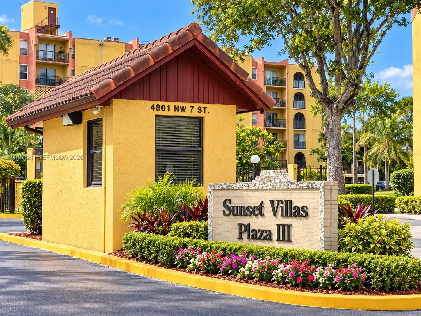 Gated community entrance featuring a guardhouse with a gabled tile roof, masonry monument signage, and mature perimeter landscaping
