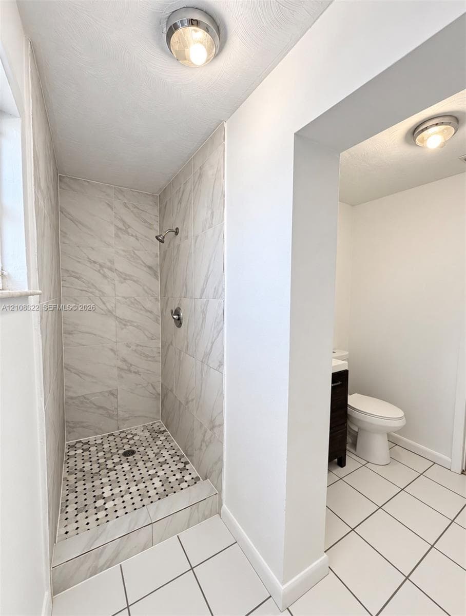 Shower stall featuring large-format light-toned stone-finish wall tile, a decorative hexagonal mosaic tile floor, and a polished metal shower fixture, paired with a separate water closet area featuring large-format white tile flooring