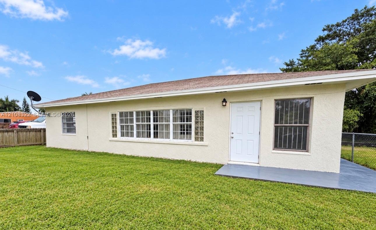 Stucco exterior featuring an expansive multi-pane window configuration, glass block accents, covered concrete patio, and mature lawn