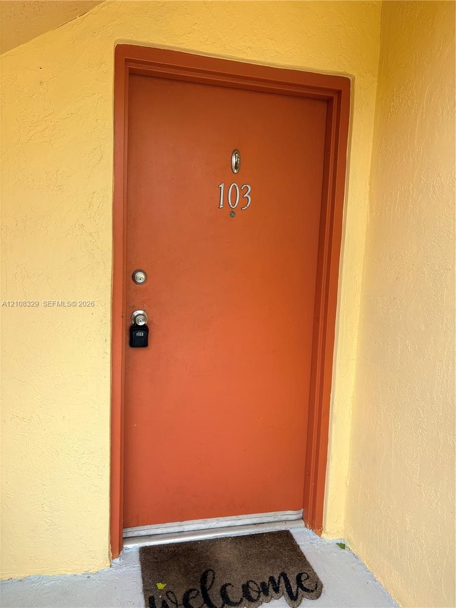 Solid orange entry door with a silver-toned door knocker and deadbolt hardware, framed by textured yellow stucco walls