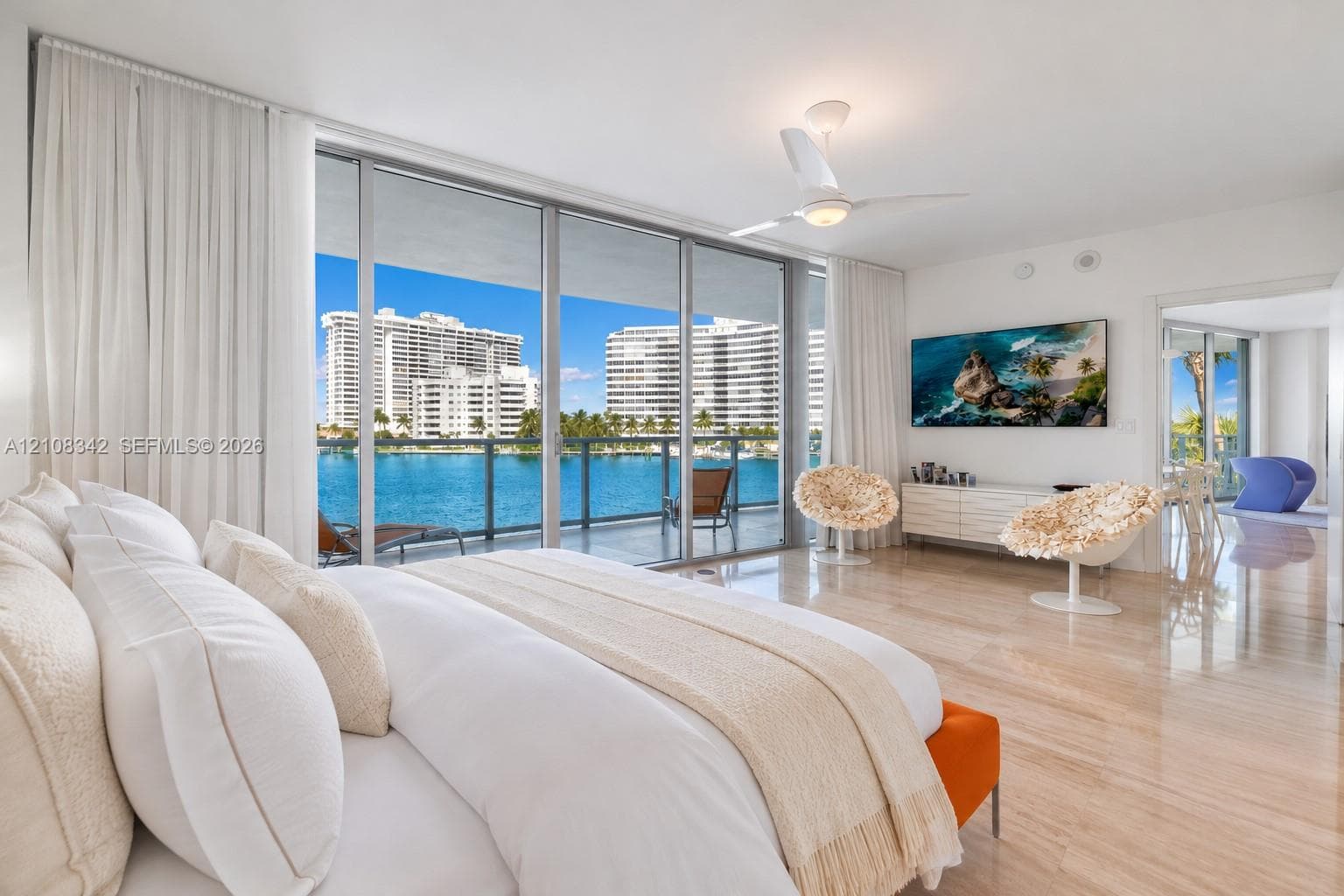 Floor-to-ceiling sliding glass doors, polished stone flooring, private glass-railed balcony, built-in ceiling fan, and prominent water backdrop