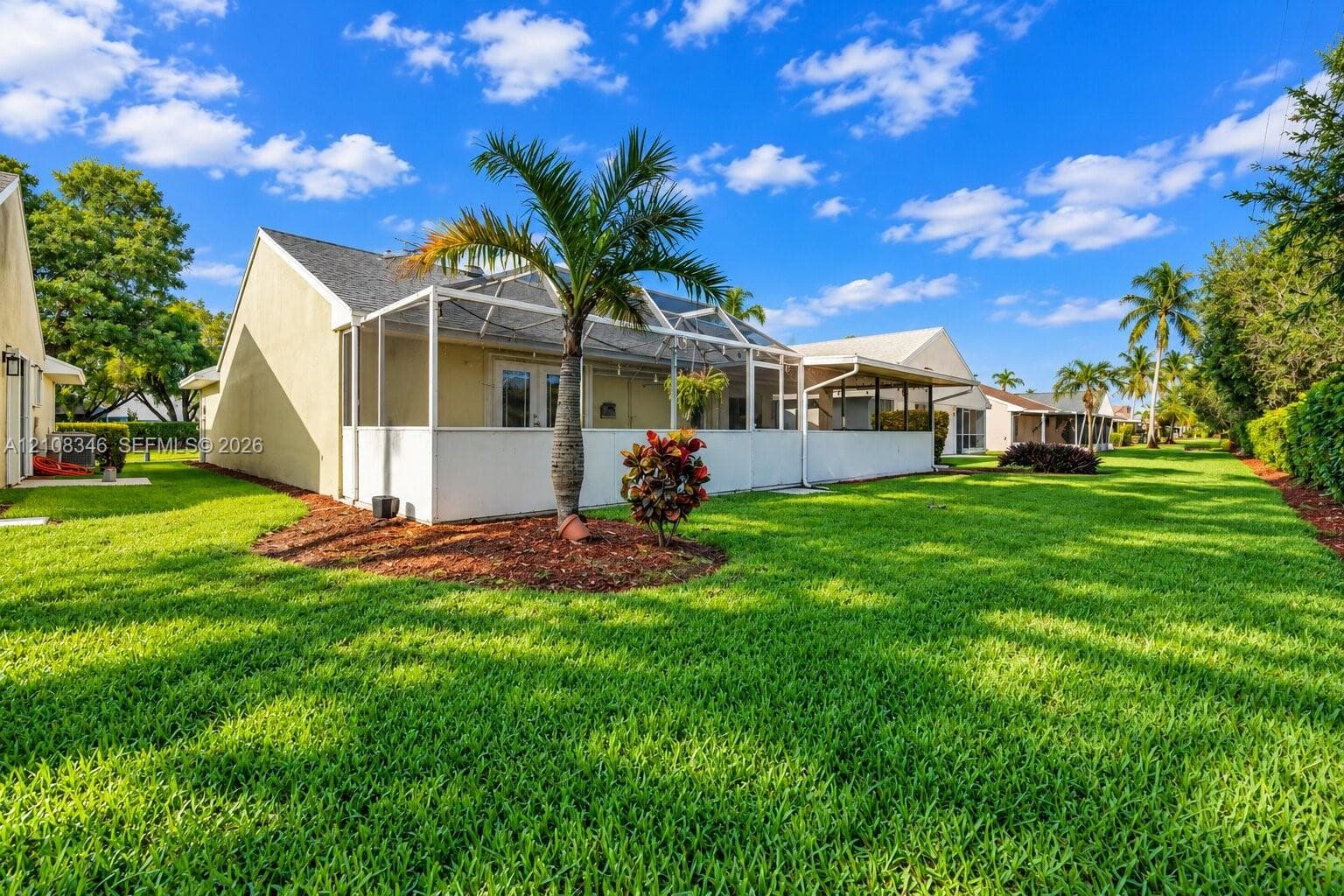 Rear exterior featuring a screened-in lanai, manicured lawn, palm trees, and ground-level plantings