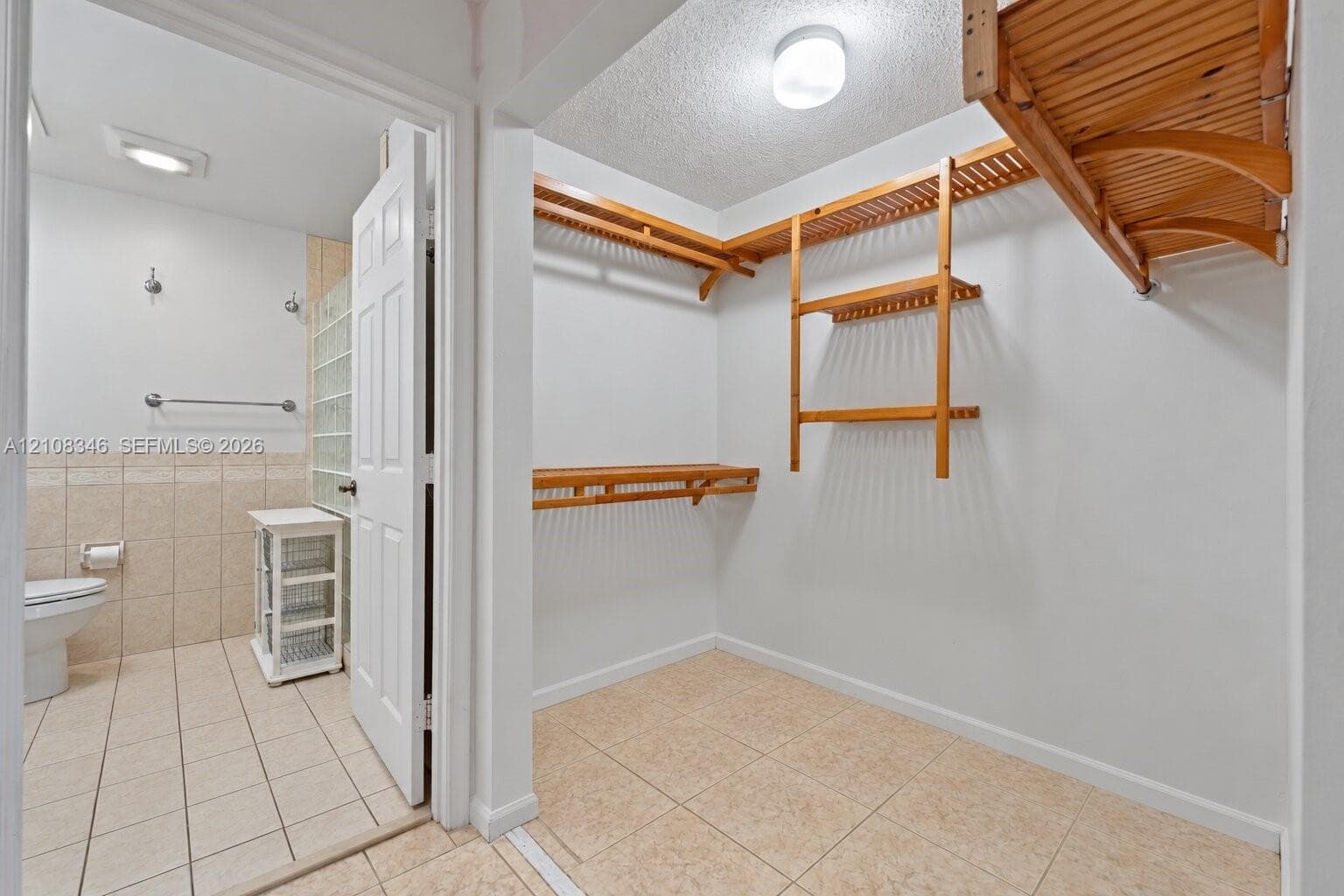 Walk-in closet featuring multi-level wooden shelving and tile flooring, with an entry to a tiled bathroom equipped with a glass block wall section and mounted hardware